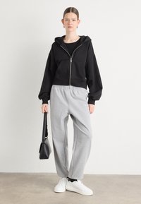Čierna mikina s kapsami na zips, kombinovaná so sivými teplákmi a bielymi teniskami. Modelka drží čiernu kabelku.