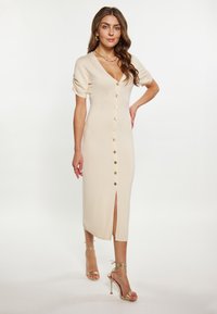 Rochie midi tricotată bej, cu decolteu în V, mâneci scurte tip puff, nasturi aurii pe față și o fanta pe coapsă. Asortată cu sandale aurii cu curele.