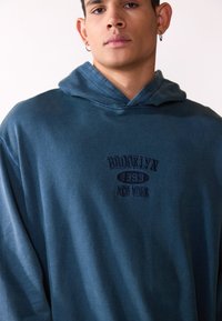 Modrá mikina s kapucňou vyrobená z mäkkého materiálu, s vyšitým textom "BROOKLYN 1993 NEW YORK" v tmavomodrej farbe. Voľný strih.