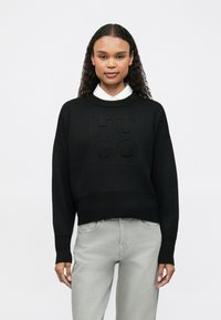 Maglione nero lavorato a maglia con logo "HUGO" testurizzato sul davanti. Ha una scollatura rotonda e maniche ampie, abbinato a jeans grigio chiaro.