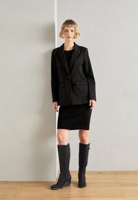 Blazer noir à revers crantés, deux poches frontales, associé à une jupe noire ajustée et des bottines compensées en suède gris montant jusqu'au genou. Fond en parquet.