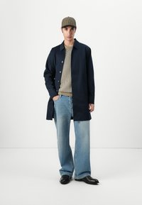 Casaco marinho com fecho de botão, usado sobre um suéter bege e jeans largos azuis. Sapatos pretos e um boné xadrez completam o visual.