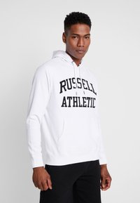 Sudadera blanca de mezcla de algodón, con un bolsillo frontal tipo canguro y un atrevido texto negro "RUSSELL ATHLETIC" impreso en el pecho.
