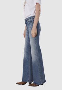 Jean flare bleu clair en denim, avec une zone déchirée, taille mi-haute et décoloration subtile. Associé à des bottes marron de style western.