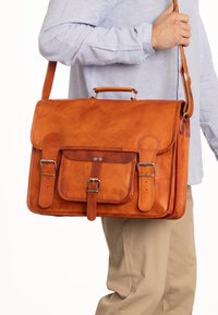 Braune Leder-Umhängetasche mit Klappe, Metallbeschlägen, Fronttasche und verstellbarem Riemen. Glatte Textur mit sichtbaren Nahtdetails.