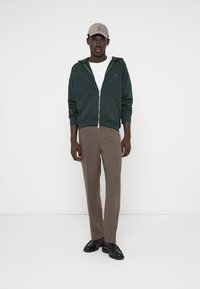 Felpa con cappuccio verde scuro con cerniera, maglietta bianca, pantaloni marroni e scarpe nere. Un cappellino grigio chiaro completa l'abbigliamento, mostrando un design casual.