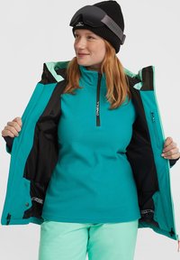 Teal fleece langmouwen laag met een halve rits, gedragen onder een zwarte en teal waterdichte jas; een zwarte gebreide beanie en skibril zijn ook zichtbaar.