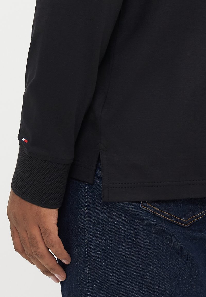 Schwarzes Langarmshirt mit texturierten Netzbündchen, seitlichen Schlitzern am Saum und einem kleinen Logo-Detail am linken Bündchen. Getragen mit blauen Jeans.