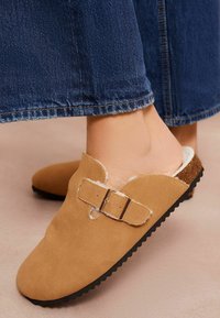Hnedé semišové klompy na slip-on so zlatou prackou, s jemným bielym podšívaním a čiernou gumovou podrážkou, čiastočne zobrazené s rozhaleným džínsovým rukávom.