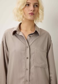 Giovane donna con capelli biondi corti, vestita con una camicia button-up beige a righe larghe, che guarda leggermente verso destra.