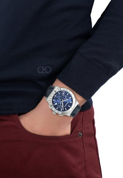 Zapešće s srebrnim kronografskim satom s plavim brojčanikom i kožnim remenom, nošen preko boje burgundca hlača i tamnoplave dugih rukava majice.