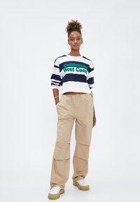 Stribet cropped sweatshirt i navy og hvid med grøn tekst "California West Coast." Beige cargo bukser med lommer, parret med kraftige hvide sko.