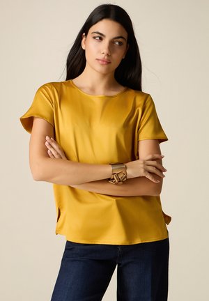 Kvinde med langt mørkt hår iført en kortærmet bluse i sennepsgul og mørke jeans, med armene krydsede, kigger til siden.