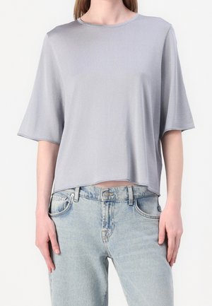 Femme portant un haut ample gris clair à manches courtes et un jean taille haute bleu clair, debout, les mains détendues le long du corps.