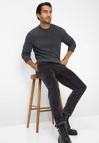 Dunkelgrauer gestrickter Pullover mit gerippten Bündchen, kombiniert mit dunklen Cordhosen und schwarzen Schnürstiefeln, sitzt auf einem Holzstuhl.