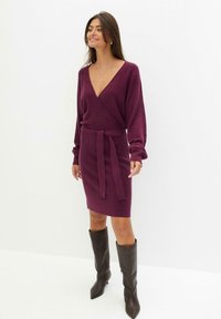 Abito avvolgente in maglia viola con scollatura a V e maniche lunghe, caratterizzato da una vita legata. Abbinato a stivali in pelle marrone alti e a punta.