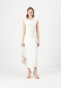 Hvit ermeløs topp med teksturerte diagonale striper, kombinert med et langt hvitt skjørt. Modellen holder en lys beige veske og har på seg nude hælsandaler.
