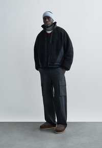 Svart jacka i konstpäls med dragkedja, lager-på-lager över en grå polo. Lösa mörka cargobyxor med fickor, bruna slip-in-skor med mönster.