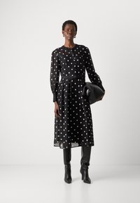Abito nero a pois con maniche lunghe, chiusura con bottoni e vita cintata. Indossato con stivali neri a punta al ginocchio e una borsa nera.