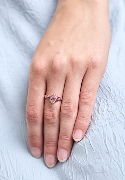 Mano con unghie naturali appoggiata su un tessuto blu chiaro, che indossa un anello d'oro con diversi piccoli gemme rosa sul dito anulare.