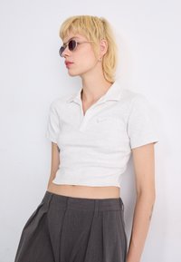 Skraćena svijetlosiva polo majica s kratkim rukavima i ovratnikom na dugme; sadrži suptilan Nike logo u bež boji. Model nosi tanke okrugle sunčane naočale.