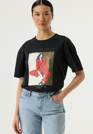 T-shirt noir en coton à manches courtes, avec une impression graphique colorée représentant une femme assise en rouge, blanc et accents dorés. Silhouette ajustée.