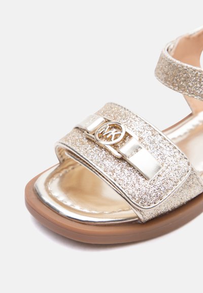 Κοντινό πλάνο λαμπερής χρυσής σαγιονάρας με καφέ σόλα και μεταλλικό λογότυπο MK στο μπροστινό λουράκι.