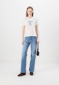 T-shirt bianco in cotone con stampa blu, abbinato a jeans in denim a campana blu. Stivaletti con tacco marroni e una piccola borsa nera. Sfondo semplice.