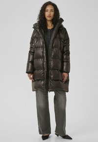 Mørkebrun dunjakke med hætte, lodret quiltede sømme og lynlås foran. Bæres over en grå top og parret med løse grå jeans og sorte sko.