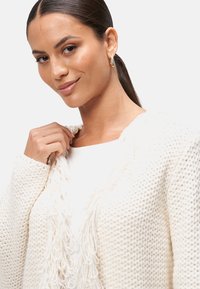 Cardigan blanc texturé avec un col à franges, porté sur un top blanc. Le tissu semble doux et épais, avec une coupe décontractée.
