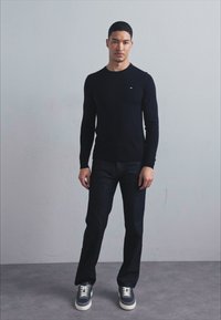Schwarzes Langarmshirt mit kleinem Logo, kombiniert mit dunklen Jeans und grauen Sneakers mit weißen Schnürsenkeln, vor einem neutralen Hintergrund stehend.
