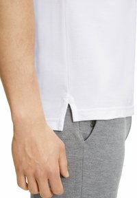 Camicetta bianca a maniche corte con una fessura laterale, abbinata a pantaloni sportivi grigi. Il tessuto della camicetta appare testurizzato e leggermente a coste.