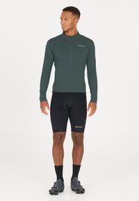 Men's long-sleeve cycling jersey in dark green, with a full zipper, paired with black padded shorts. Lightweight, breathable fabric, and fitted design.