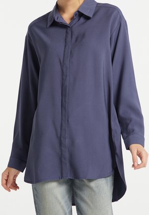 Personne portant une chemise bleu foncé ample à manches longues avec des boutons, un ourlet plus court devant et plus long derrière, et un jean clair visible en bas.