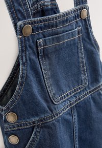 Primo piano di salopette in denim blu scuro con tasca frontale sul petto e bottoni metallici su spalline e lati.