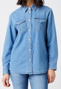 Light blue denim shirt with long sleeves, button-up front, and two patch pockets. Features subtle western-style yoke and stitching details.