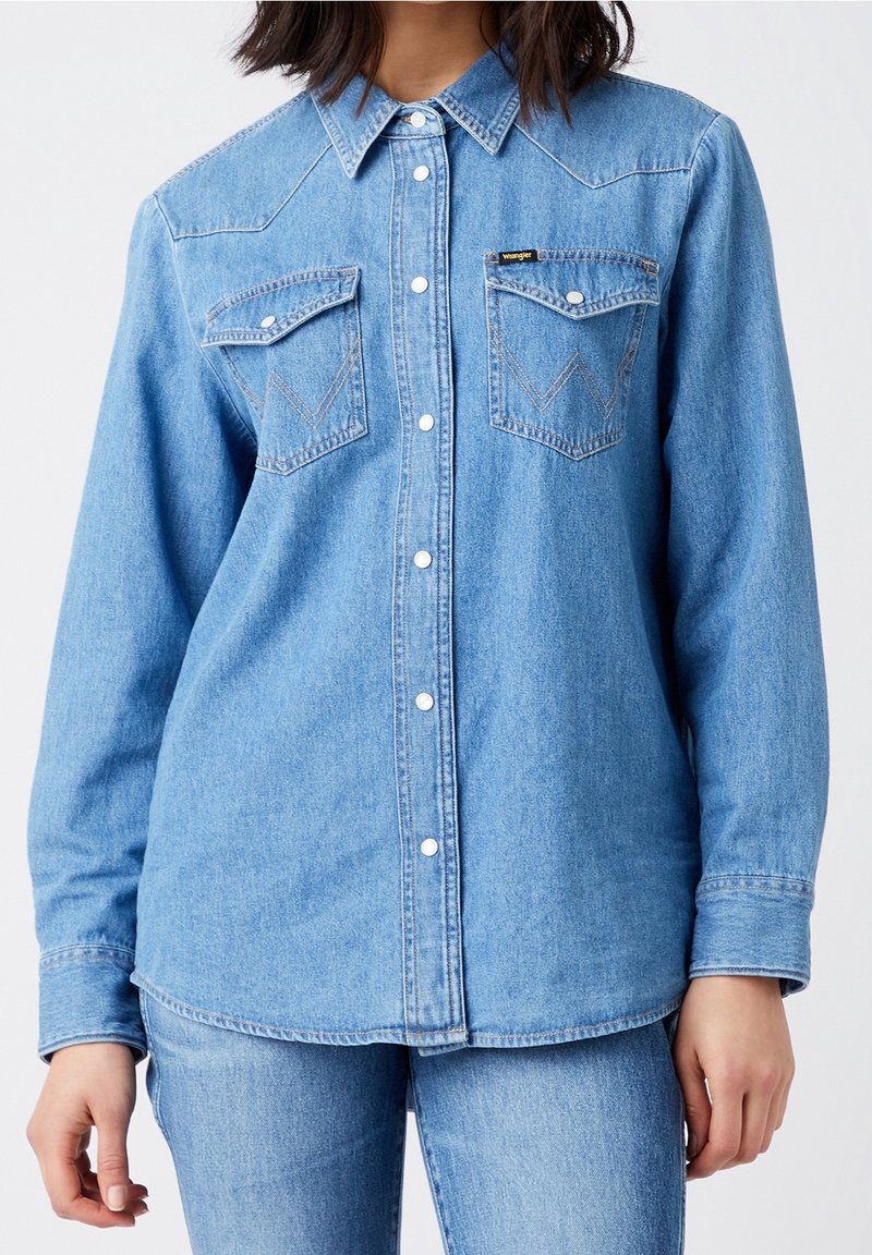 Light blue denim shirt with long sleeves, button-up front, and two patch pockets. Features subtle western-style yoke and stitching details.