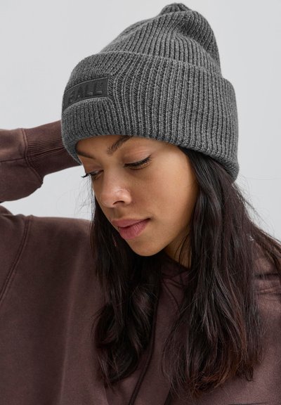 Sieviete ar tumšiem matiem, ģērbusies pelēkā ribotā adīšanas cepurē un brūnā kapucē, skatās uz leju ar vienu roku paceltu pie galvas aizmugures.