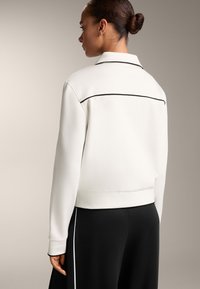 Fehér pulóver gallérral, fekete díszítéssel a vállakon és bordázott mandzsettával, fekete nadrággal kombinálva. Simaság és hétköznapi stílus jellemzi.