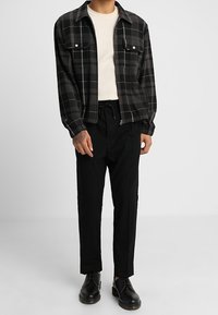 Giacca a quadri nera e grigia con due tasche sul petto, abbinata a un maglione a coste crema e pantaloni neri a sigaretta. Scarpe nere completano l'outfit.