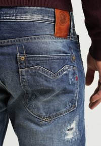 Jeans de mezclilla con un lavado azul desgastado, detalles desgastados y un parche de cuero en la cintura. Presenta un patrón de costura de bolsillo distintivo.