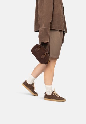 Braune Wildleder-Handtasche mit goldenen Beschlägen, getragen beim Gehen. Kombiniert mit einer braunen Jacke, Shorts und braunen Sneakers mit Gummisohle.