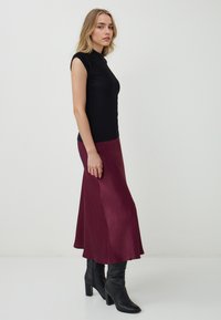 Top senza maniche nero abbinato a una gonna midi di raso bordeaux con orlo svasato, e stivali neri al ginocchio con tacco a design morbido.