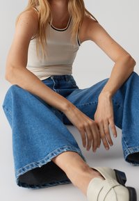 Henkilö, joka on pukeutunut valkoiseen hihattomaan ribbipaitaan, leveisiin sinisiin farkkuihin ja valkoisiin liukuvaihtoehtoisiin kenkiin, istuu lattialla yksinkertaisen taustan edessä.