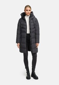 Dunjacka i mörkgrått, lång design med huva, fyrkantig quiltad mönster och dragkedja framtill. Bärs över en vit polo och svarta jeans.