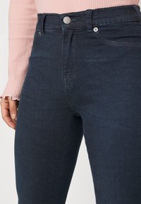 Jeans de mezclilla oscuros de cintura alta con una textura suave, que cuentan con un cierre de botón metálico y costuras sutiles a lo largo de los bolsillos.