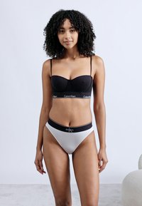 Νεαρή γυναίκα με σγουρά μαλλιά που φοράει μαύρο σουτιέν Calvin Klein και άσπρο εσώρουχο Calvin Klein, στέκεται μπροστά σε απλό φωτεινό φόντο.