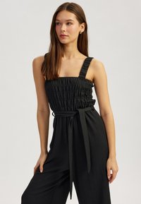 Mono negro hecho de tela texturizada con tirantes anchos, cuerpo fruncido y un detalle de cintura atada. El diseño presenta un ajuste relajado.