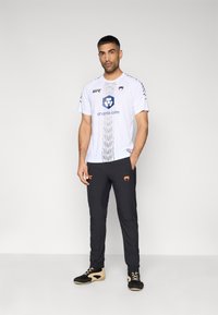 Λευκό αθλητικό πουκάμισο με μαύρες λεπτομέρειες, με το λογότυπο UFC και μεγάλο γραφικό σχέδιο, σε συνδυασμό με μαύρο παντελόνι και μπεζ αθλητικά παπούτσια.