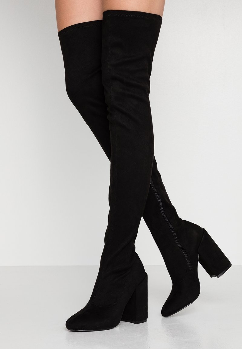 Bottes noires en daim au-dessus du genou avec talons carrés portées sur des jambes croisées sur un fond clair uni.